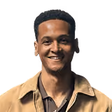Smiling man with short curly hair wearing a tan jacket over a dark shirt.
