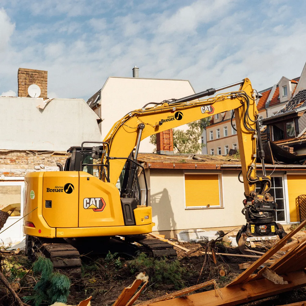 Gelber CAT-Bagger mit Abriss Breuer-Logo inmitten von Baustellenschutt vor teilweise abgerissenem Gebäude in Berlin