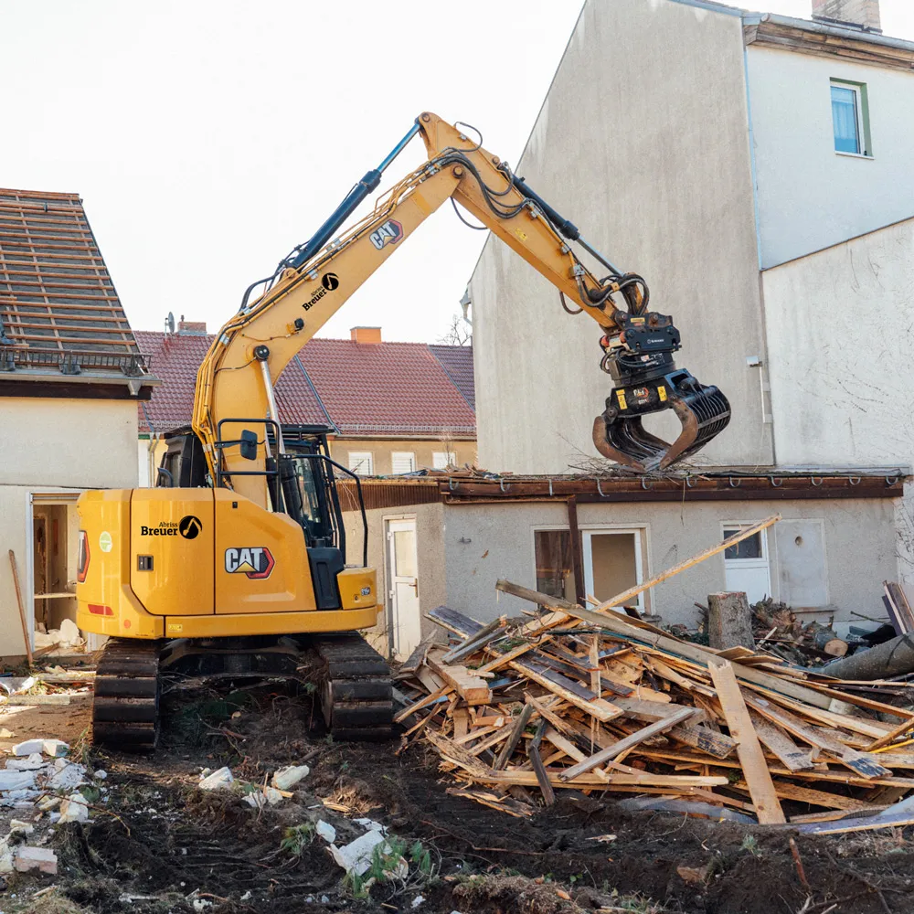 Gelber Bagger mit Greifarm räumt Holz- und Baumaterialien auf Baustelle neben Wohngebäuden in Berlin
