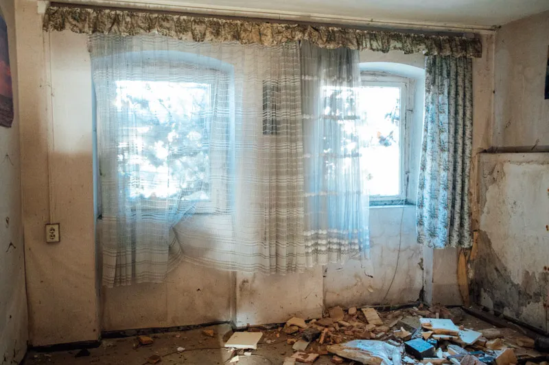 Entrümpelung Brandenburg: Verlassenes Zimmer mit zwei Fenstern, halbtransparenten Gardinen und zerbrochenem Mauerwerk auf dem Boden.