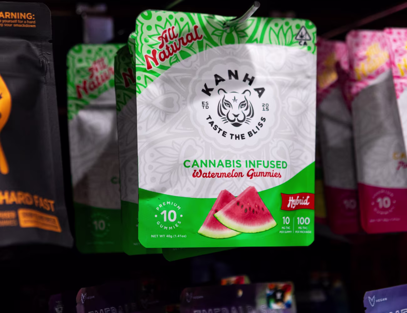 Close-up of a Kanha cannabis-infused watermelon gummies package on a dispensary shelf, labeled hybrid with 10 gummies and 100mg THC per package, surrounded by other colorful edible products in the background.