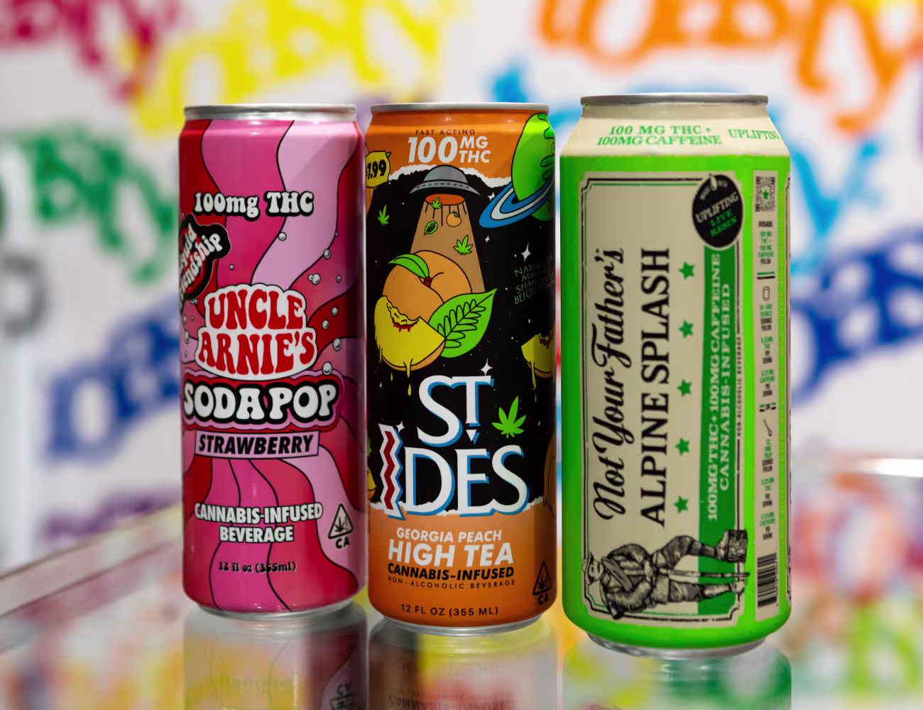  Three colorful cannabis-infused beverage cans displayed side by side, including Uncle Arnie’s Strawberry Soda Pop, ST Ides Georgia Peach High Tea, and Alpine Splash, each labeled with 100mg THC, set against a vibrant graffiti-style background.