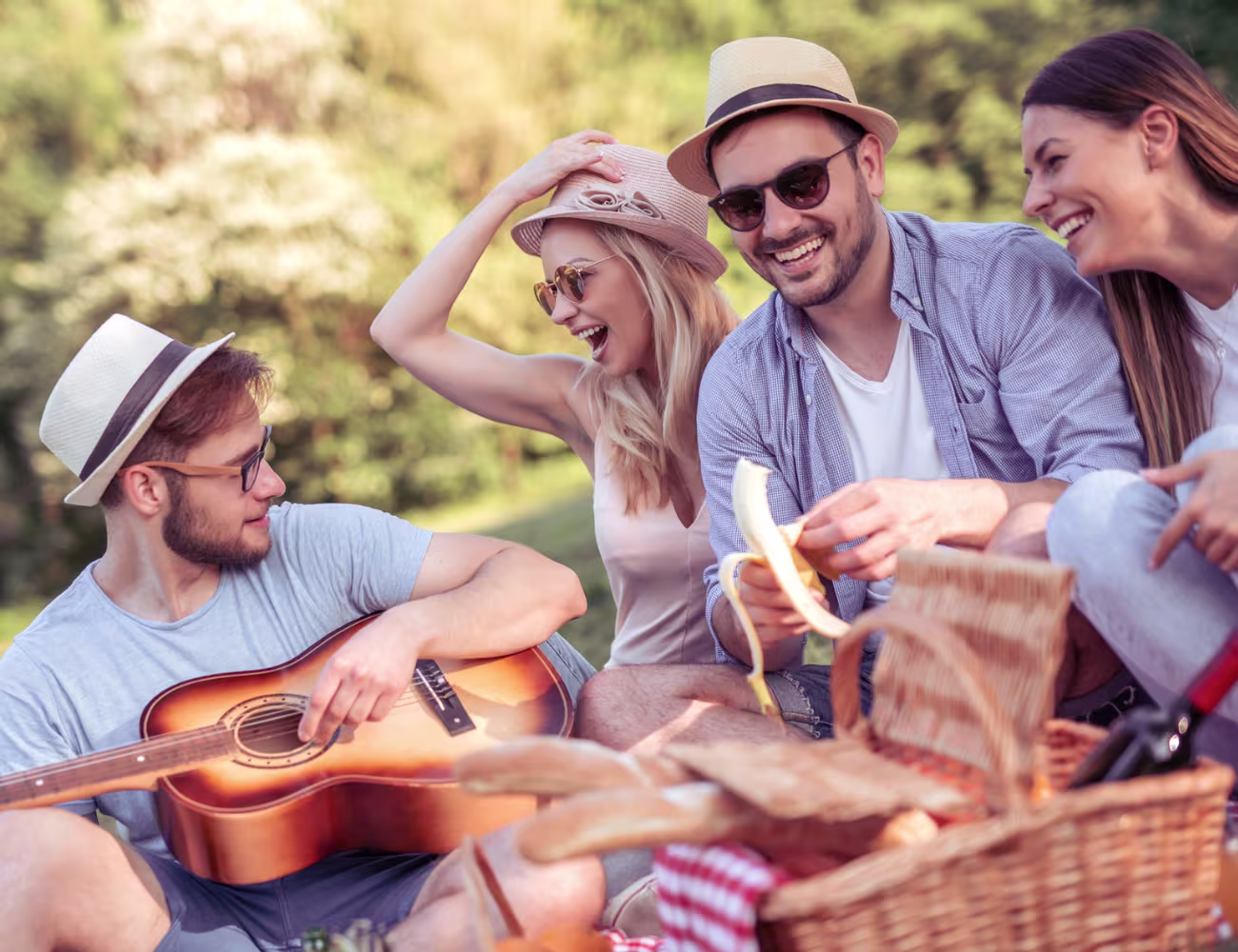Group of friends enjoying a sunny outdoor picnic, laughing together with a guitar, snacks, and drinks in a relaxed park setting.