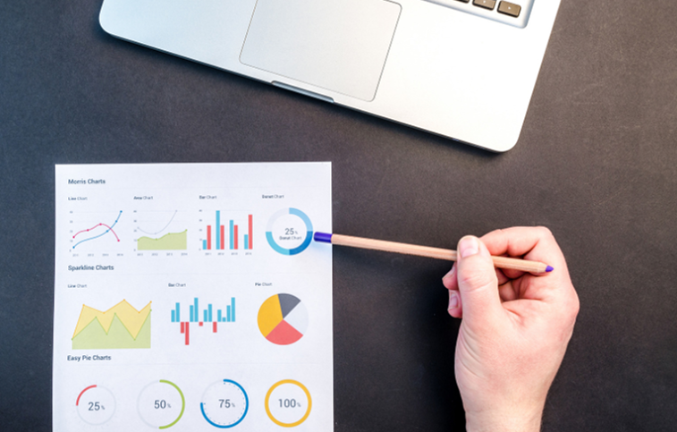 Businessperson pointing at printed analytics charts on a desk next to a laptop. i-radius