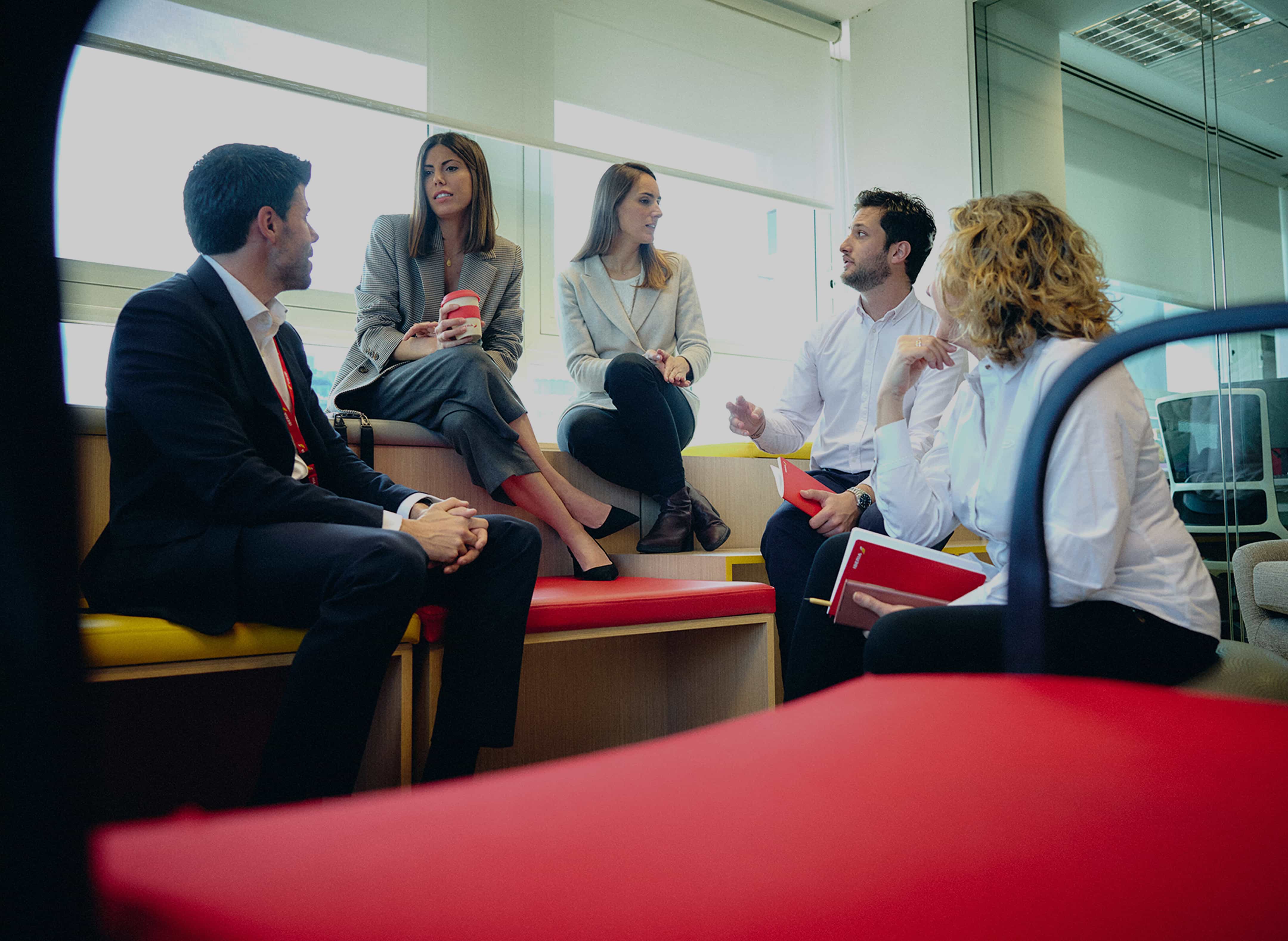 Cinco profesionales conversando en una sala de reuniones moderna con ventanales, sentados en bancos de madera con cojines rojos y amarillos, algunos con cuadernos y una taza de café.