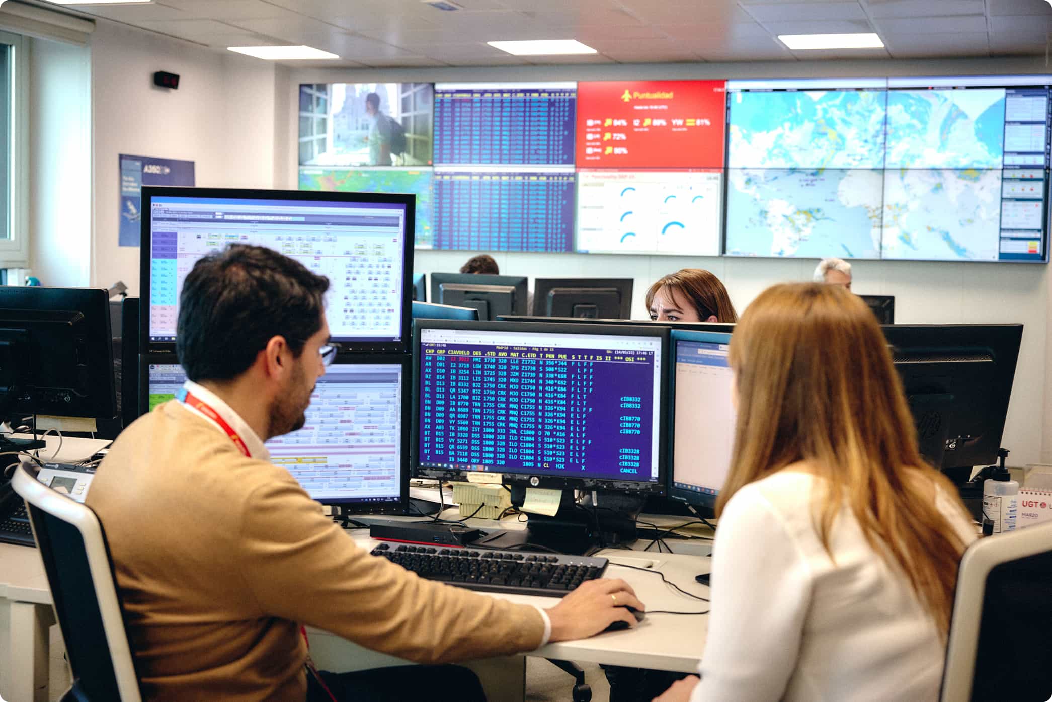Oficina con varias personas trabajando en escritorios con múltiples pantallas de computadora mostrando datos y mapas, y una pared con una gran pantalla múltiple que muestra información diversa, incluyendo mapas y estadísticas de vuelos.