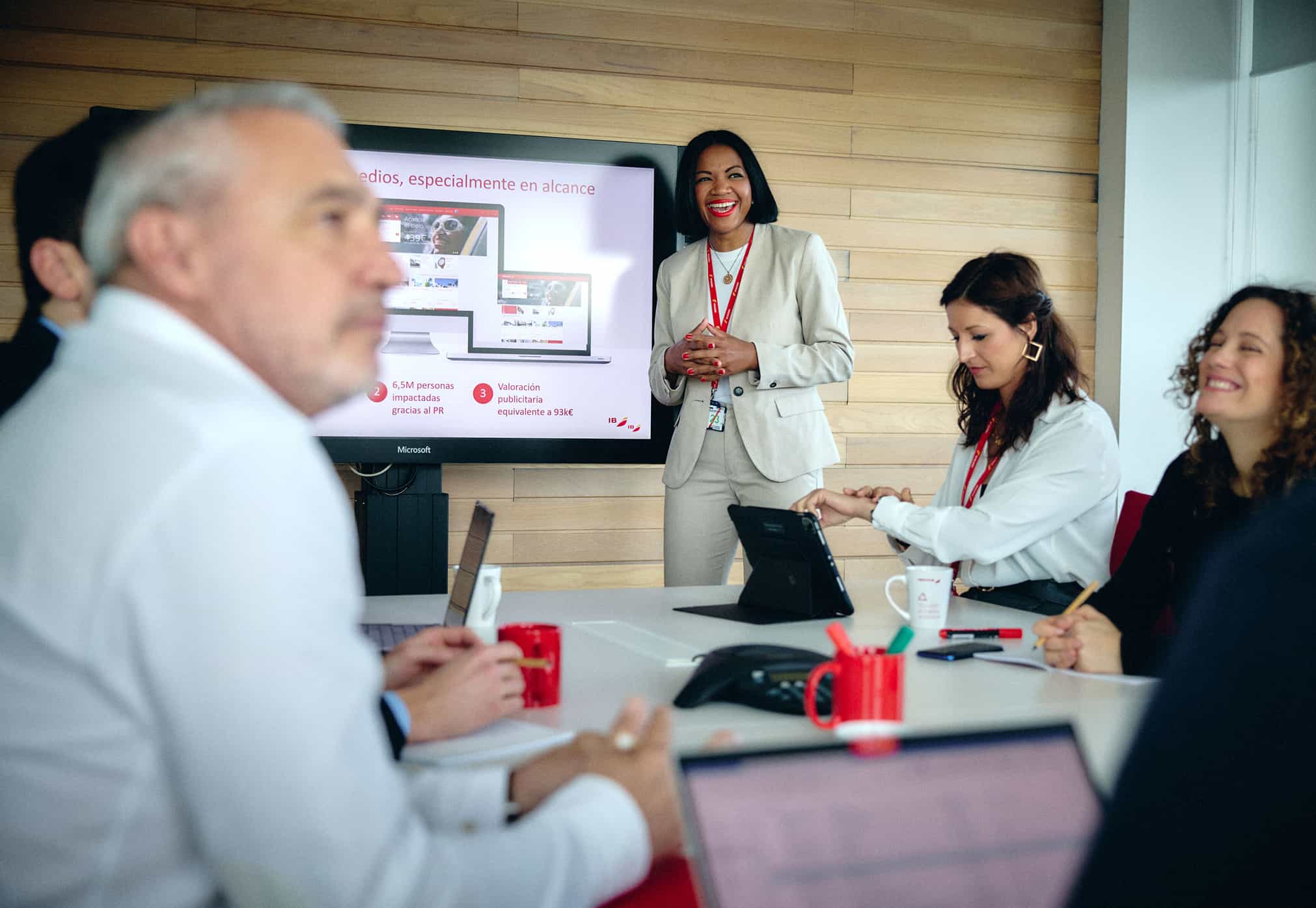 Mujer sonriente con traje beige presenta información en una pantalla durante una reunión de trabajo en una sala con paredes de madera, mientras varias personas escuchan y toman notas.
