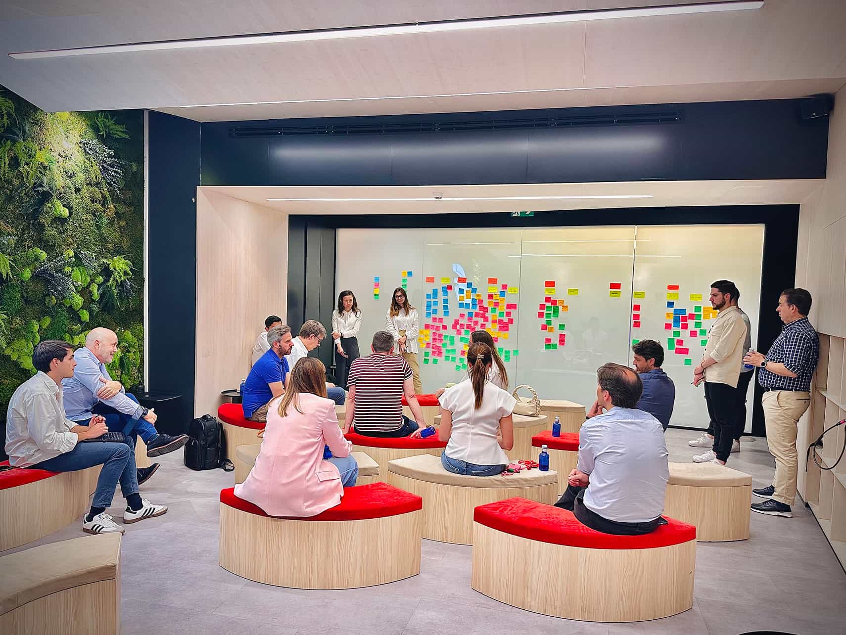 Grupo de personas en una reunión o sesión de trabajo en una sala moderna con asientos semicirculares de madera con cojines rojos, frente a una pizarra de vidrio llena de notas adhesivas de colores organizadas en columnas, mientras dos mujeres de pie explican o presentan algo al grupo. A la izquierda hay una pared con vegetación y todos parecen concentrados en la actividad.