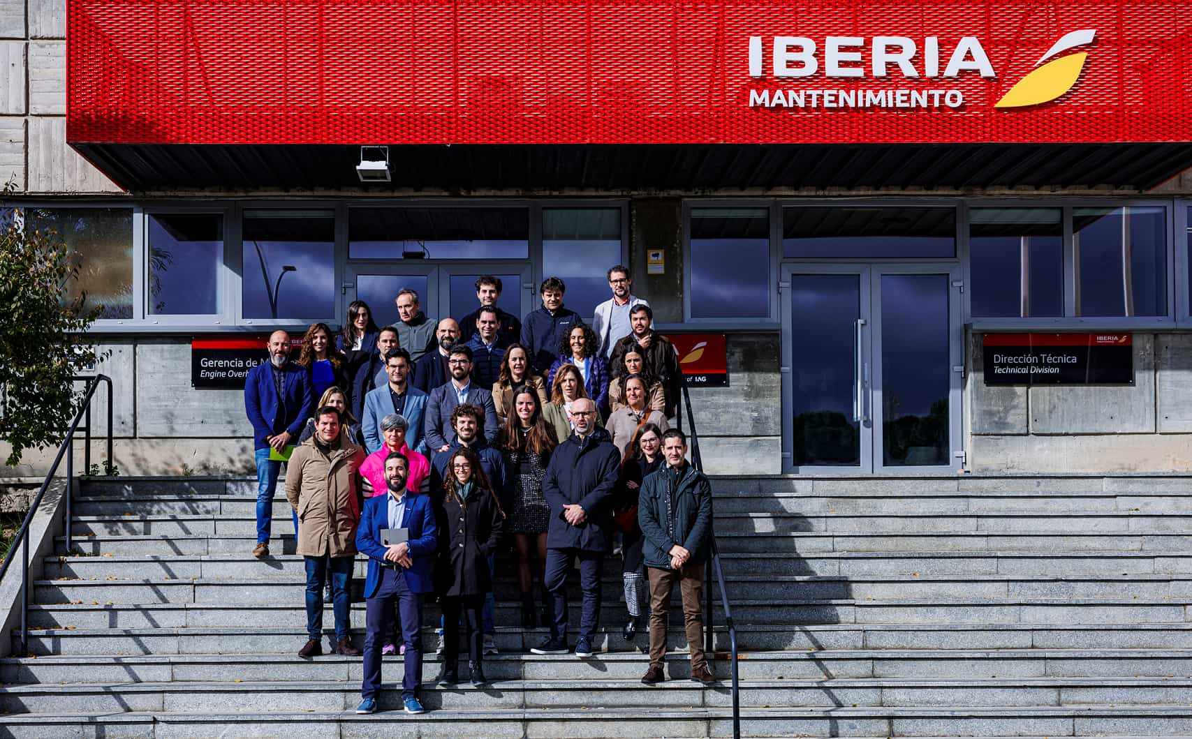 Un grupo de personas posando en las escaleras frente a un edificio con un cartel rojo que dice Iberia Mantenimiento, con placas que indican Gerencia de Motores y Dirección Técnica.