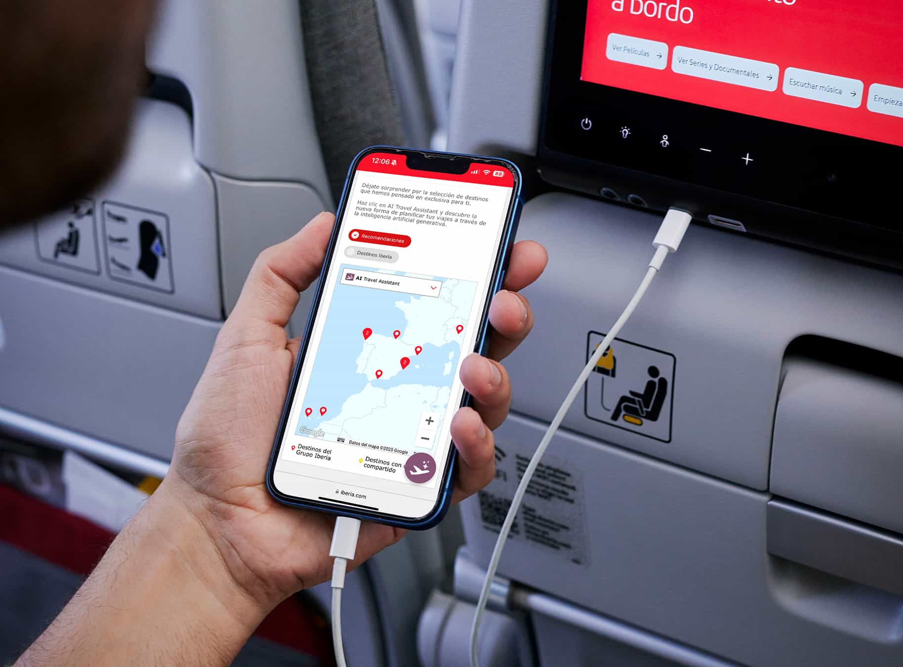 Una mano sostiene un teléfono móvil que muestra una aplicación de asistente de viaje de IA con un mapa y destinos marcados, mientras está conectado con un cable USB a una pantalla de entretenimiento en un asiento de avión.