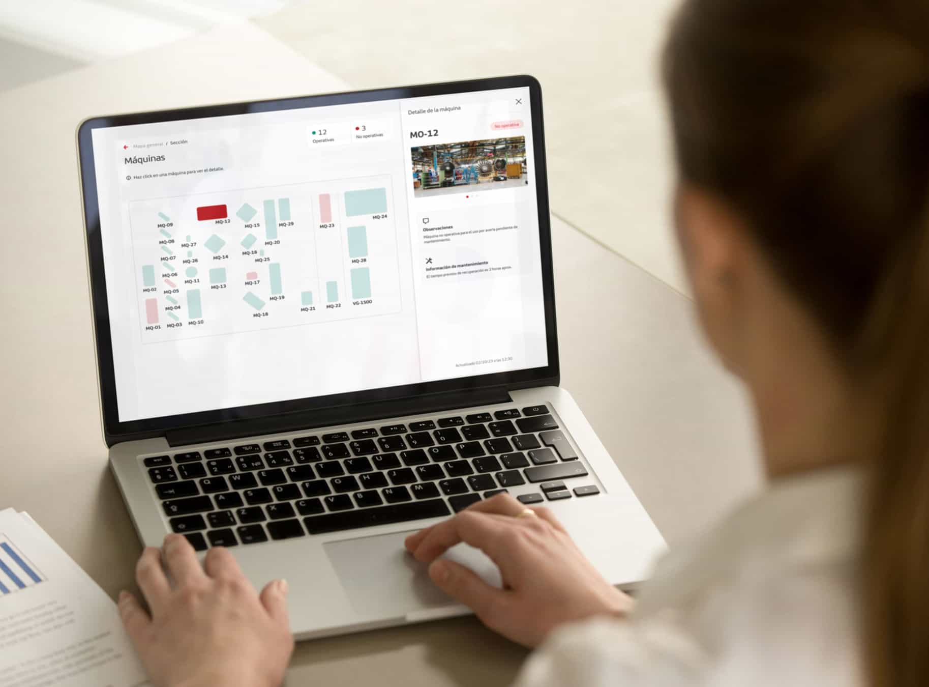 Persona usando una computadora portátil que muestra un panel de control de máquinas con un mapa de máquinas identificadas por códigos y un cuadro de detalles de la máquina MO-12, con indicadores de operativas y no operativas.