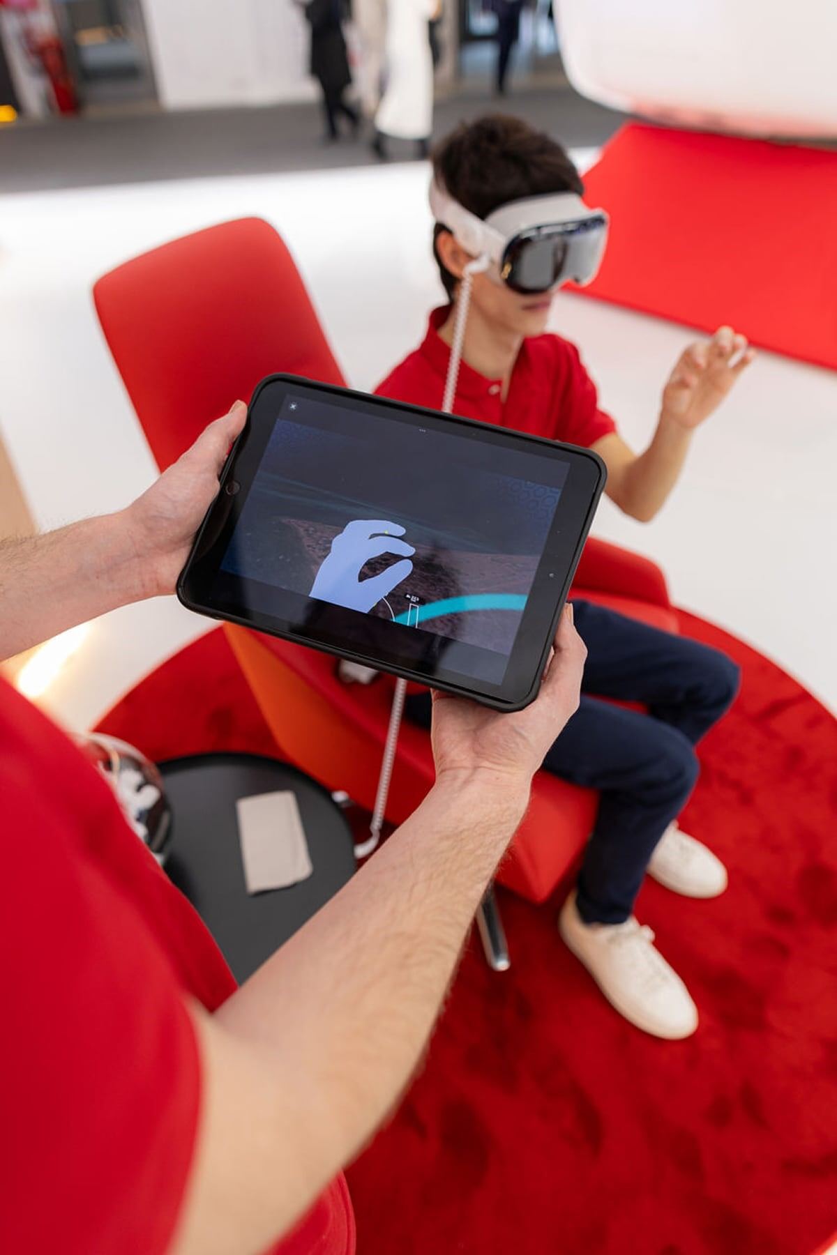 Un niño con una camiseta roja y pantalones oscuros usa gafas de realidad virtual sentado en una silla roja sobre una alfombra roja, mientras una persona sostiene una tableta que muestra una mano virtual replicando los movimientos del niño.