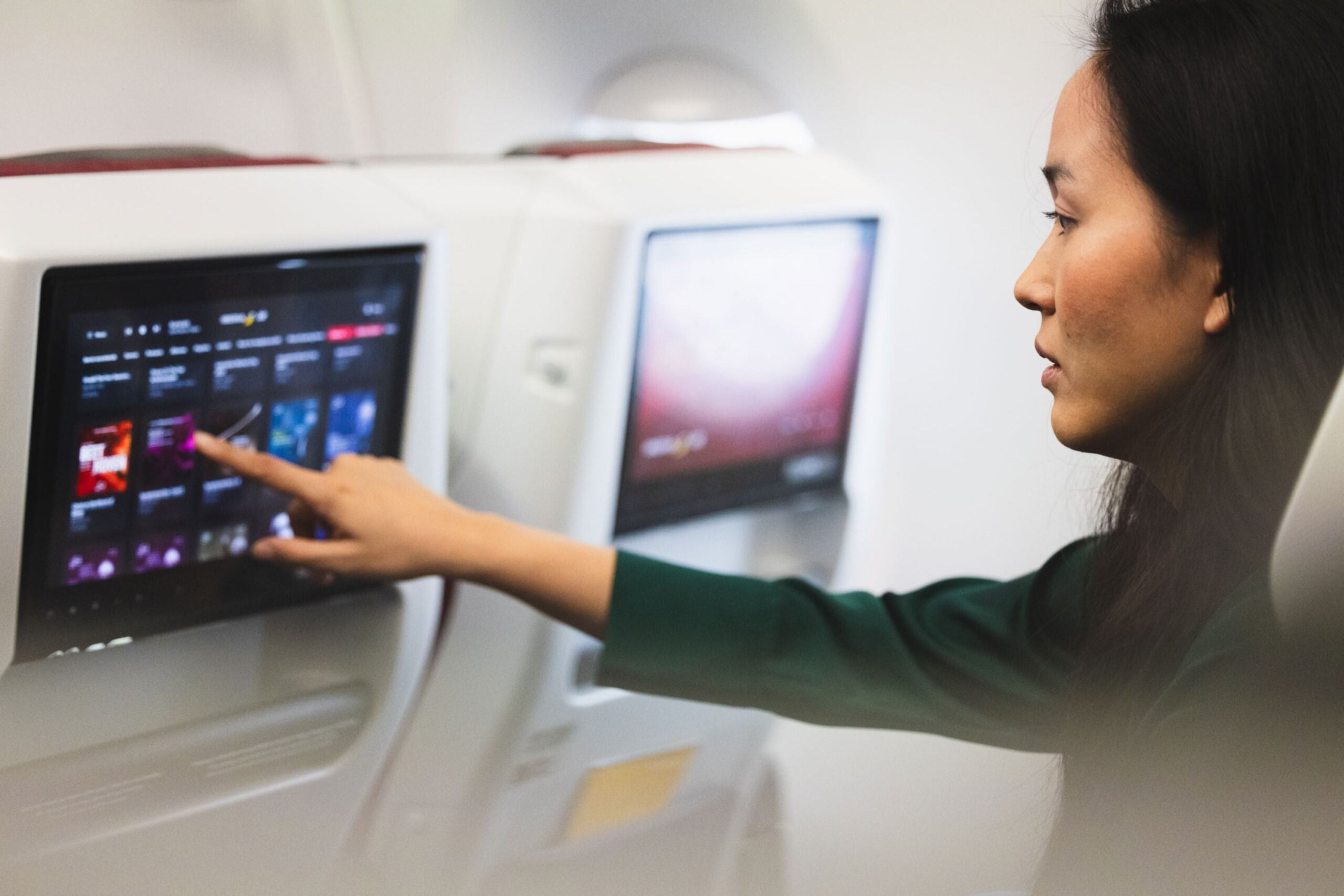 Mujer con cabello largo y oscuro tocando la pantalla táctil de entretenimiento en el respaldo de un asiento dentro de un avión.
