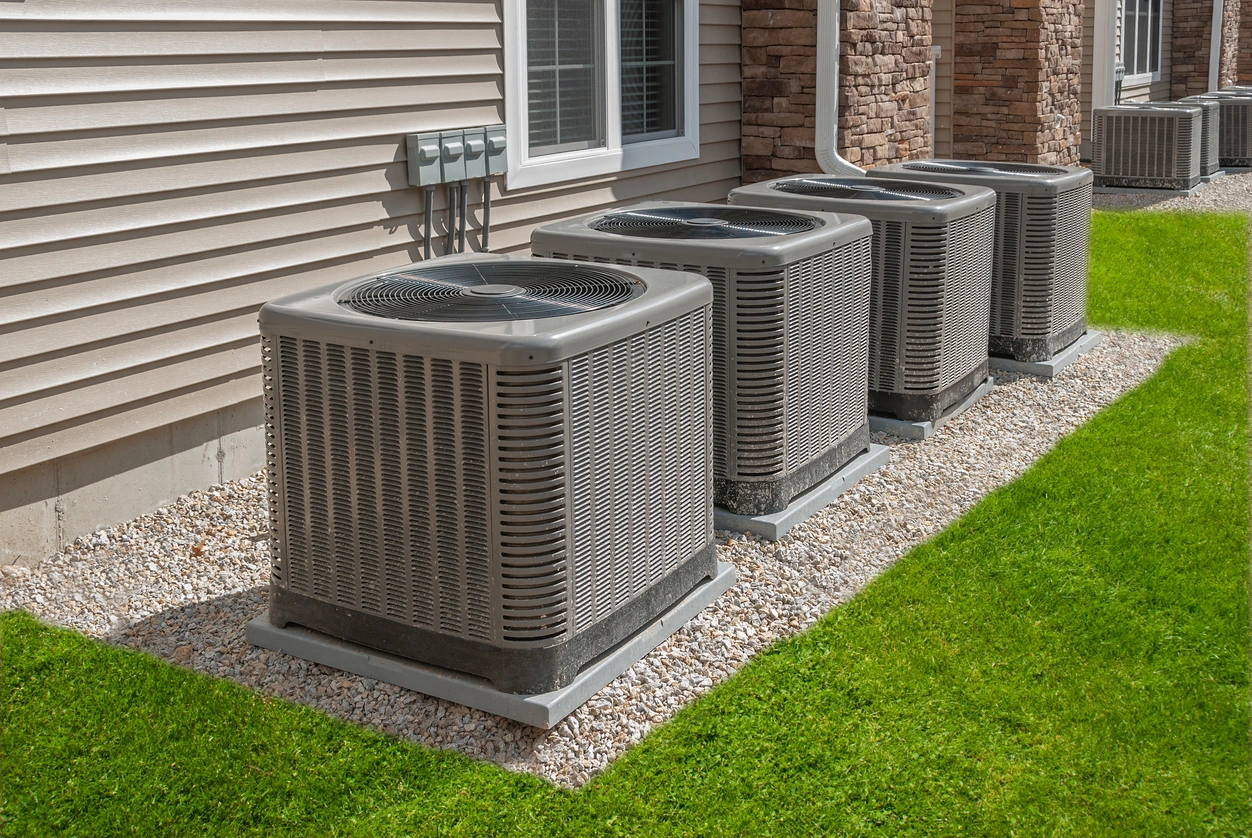 Four outdoor air conditioning units on gravel beside a house with beige siding.
