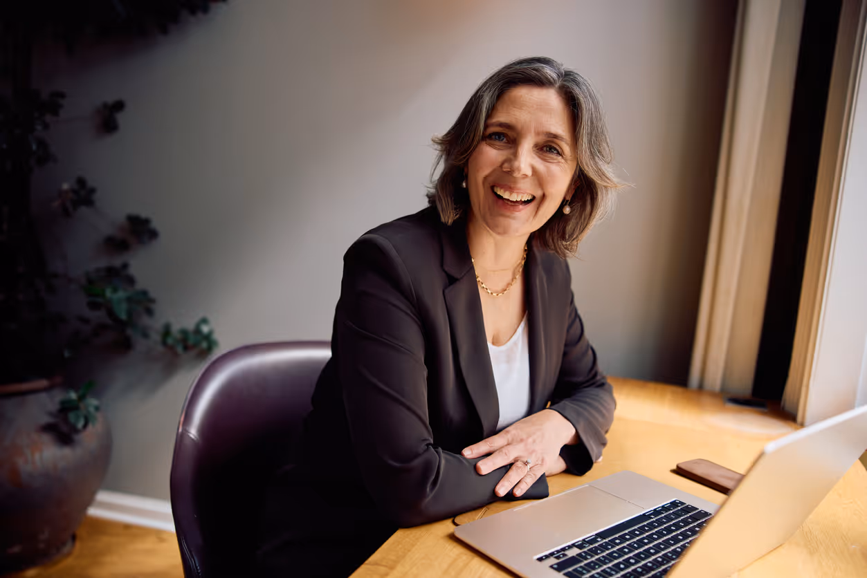 Smiling middle-aged woman in a black blazer sitting at a wooden desk with a laptop and looking at the camera.