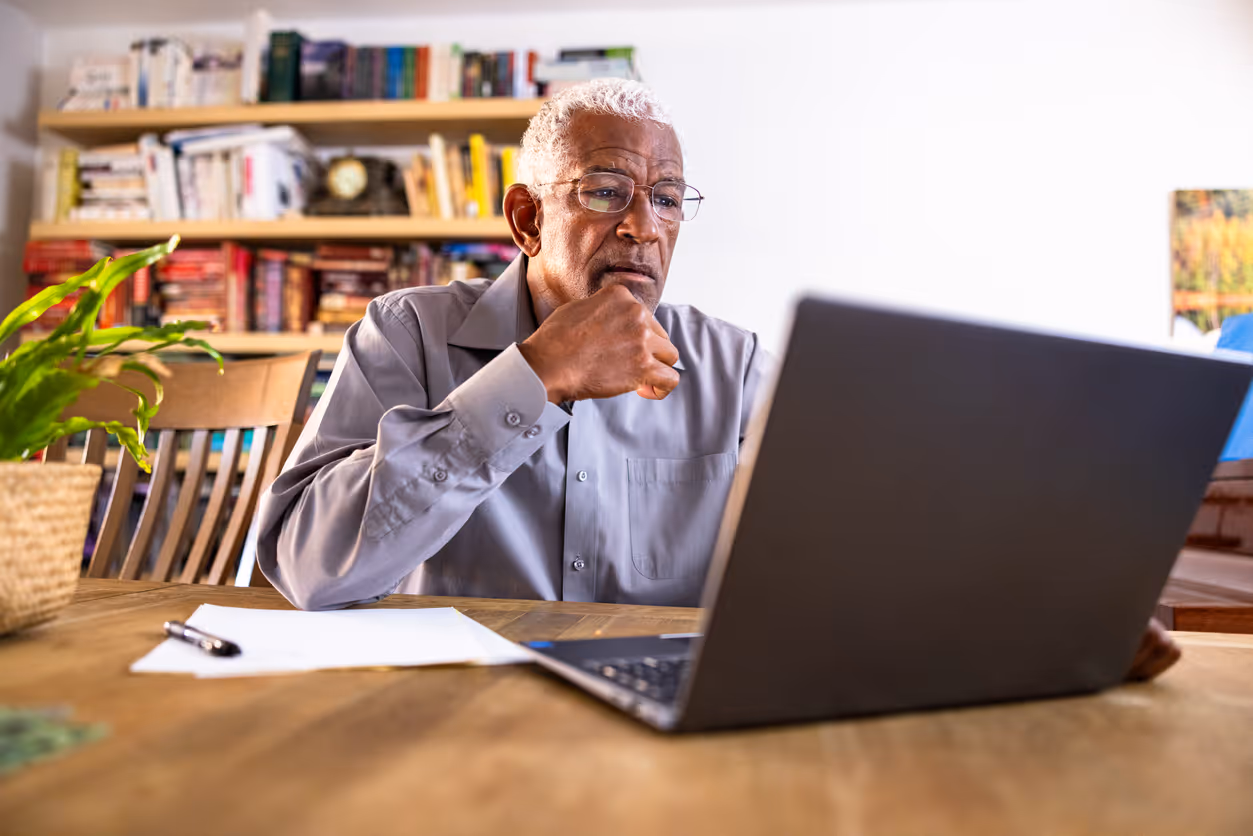 Elderly man wearing glasses and a gray shirt thoughtfully looking at a laptop while sitting at a wooden table with papers and a pen, in a room with bookshelves.