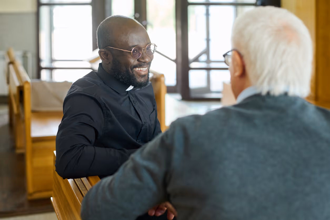 Smiling African American priest wearing glasses and a clerical collar talking to an older man with white hair inside a church.