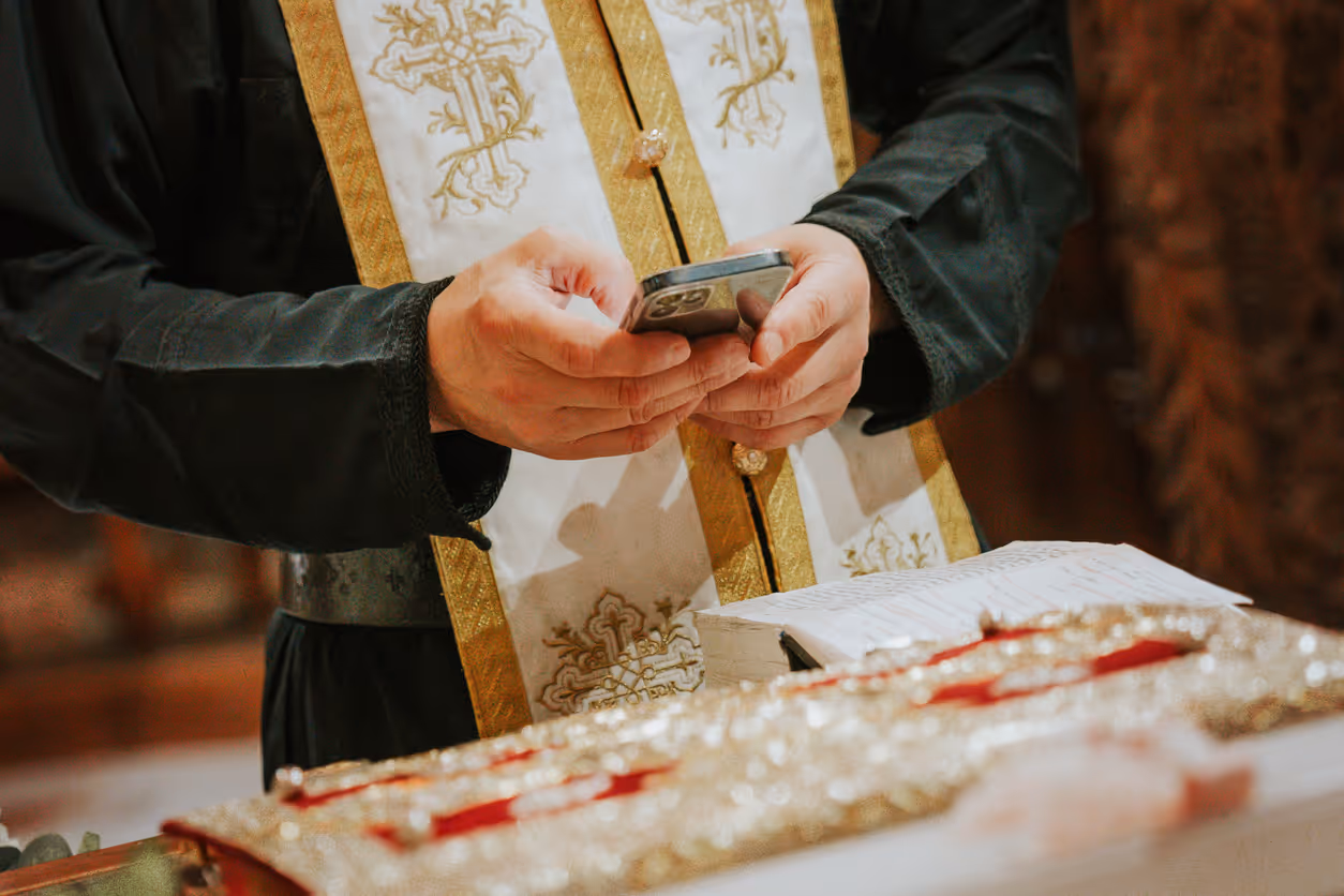 Clergyman in black robe with white and gold embroidered stole holding a smartphone over an open religious book.