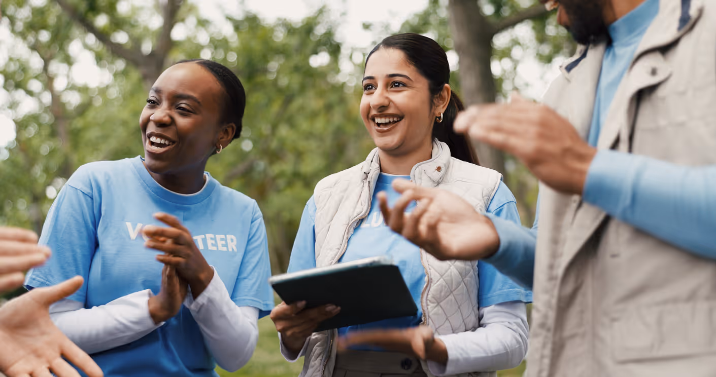 Smiling diverse volunteers in blue t-shirts clapping and holding a tablet outdoors in a park.