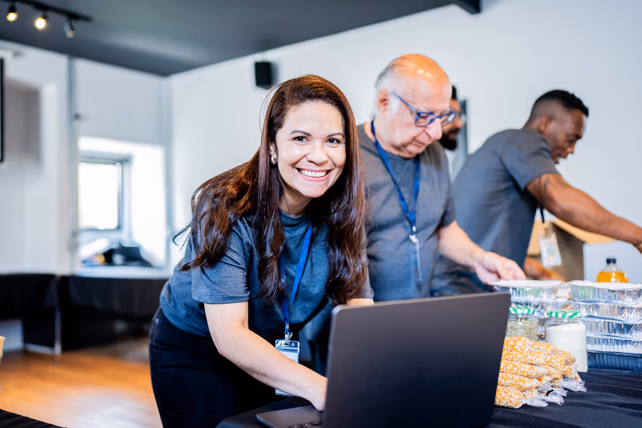 Smiling woman working on a laptop with two men organizing packaged food on a table in a bright room.