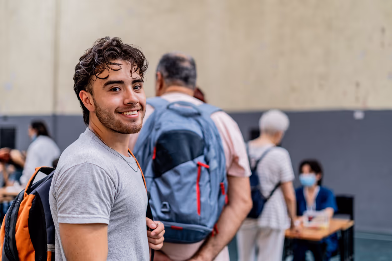 Young man with a backpack smiling at the camera in an indoor setting with people in line in the background.