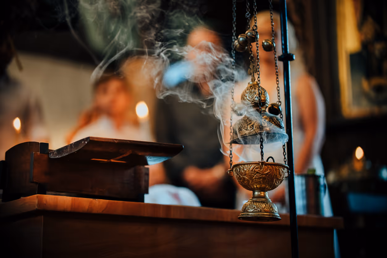 Ornate brass incense burner hanging and emitting smoke, with blurred people and candles in the background.
