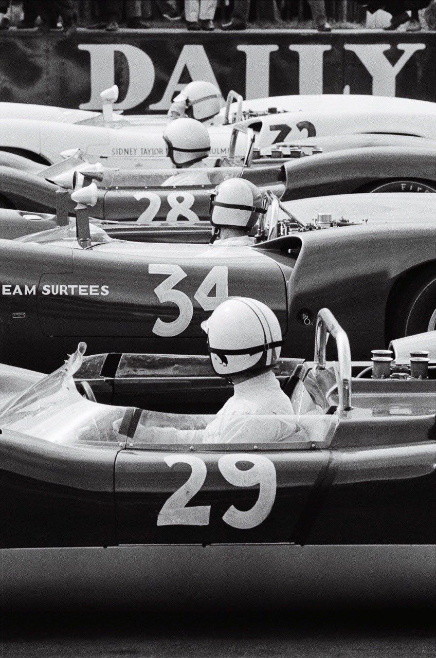 Four vintage race cars with numbers 29, 34, 28, and 72, each driven by a helmeted driver, lined up closely on a racetrack before a race.