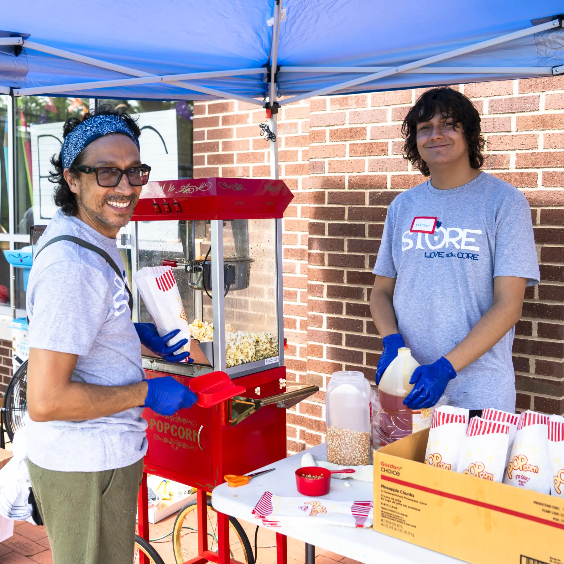 A volunteer is operating a popcorn machine and poses with a community member holding a fresh bag of popcorn