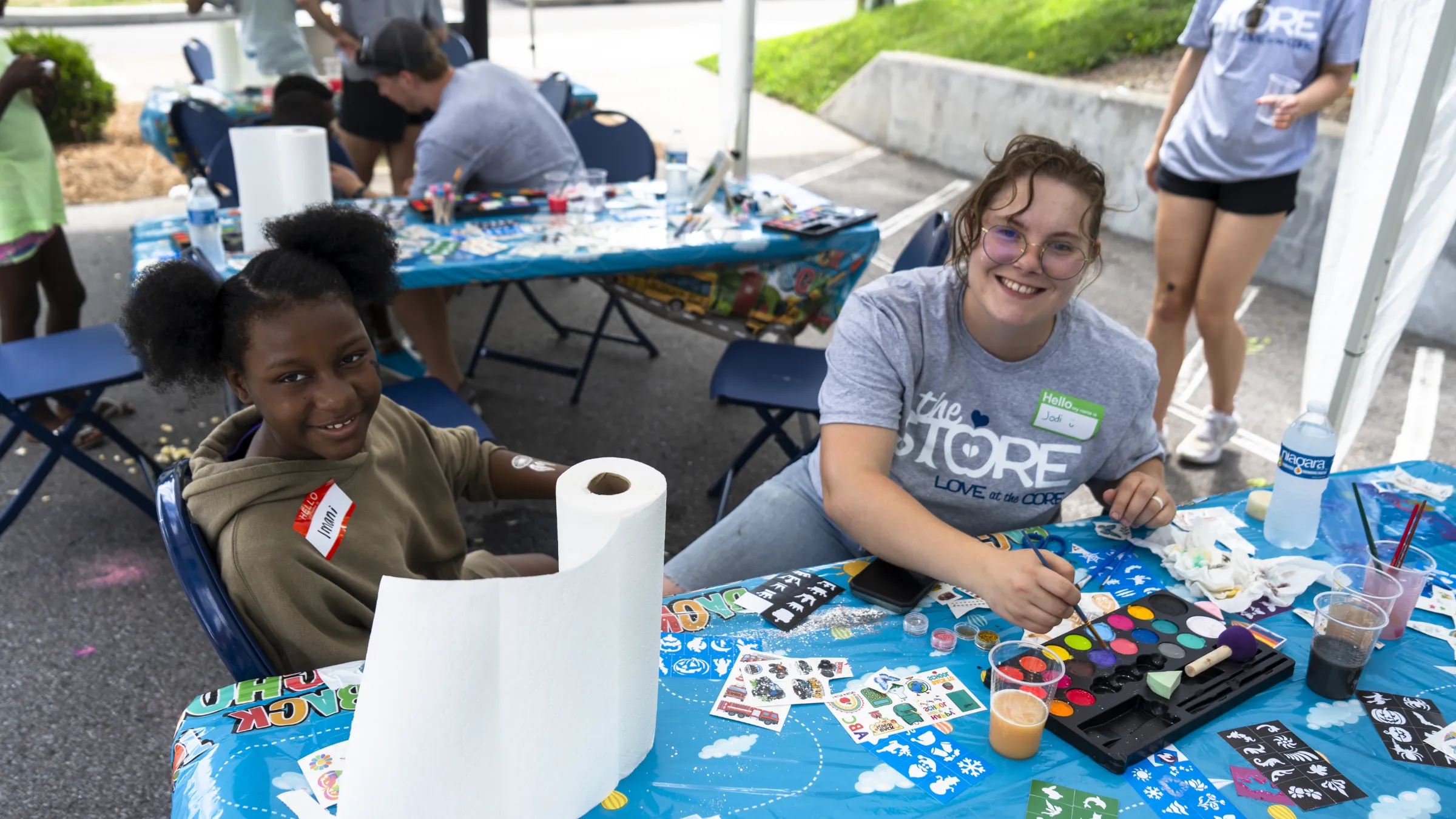 A volunteer sits at a crafts table with a young girl from the community making crafts with more tables and community members behind them.