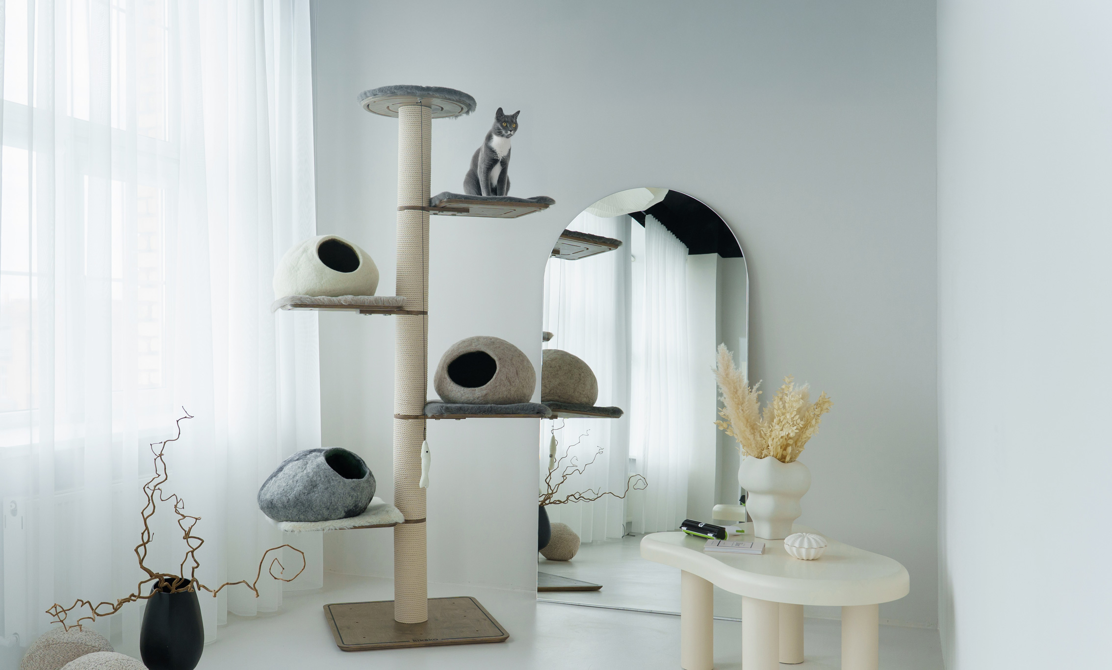 Tall Kikska cat tree with wool caves and a grey cat on the top platform in a bright white room