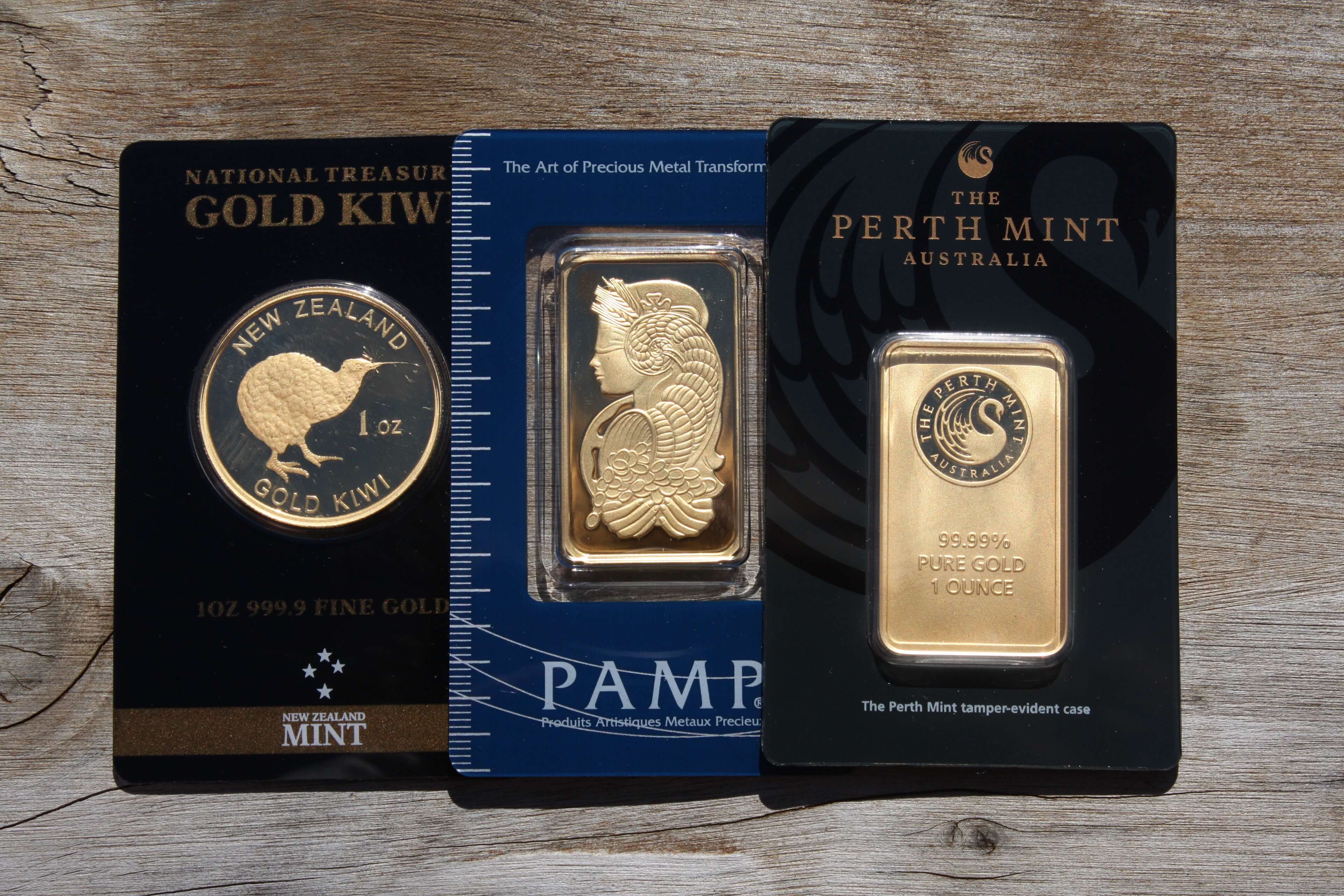 Three packaged gold bars and coins from New Zealand Mint, PAMP, and Perth Mint arranged on a wooden surface updated.