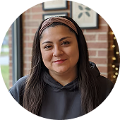 Mujer joven con cabello largo y lacio, usando una sudadera negra y una diadema, sonriendo con fondo de muro de ladrillos y ventanas.