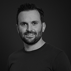 Retrato en blanco y negro de un hombre con barba y cabello corto, vestido con camiseta negra, sonriendo ligeramente frente a fondo oscuro.