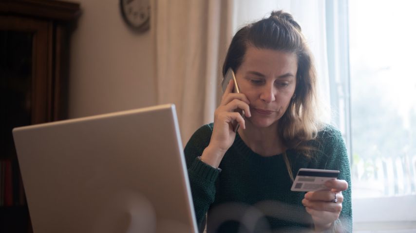 Mujer preocupada hablando por teléfono mientras sostiene una tarjeta bancaria frente a una computadora portátil, representando una posible estafa por transferencia.