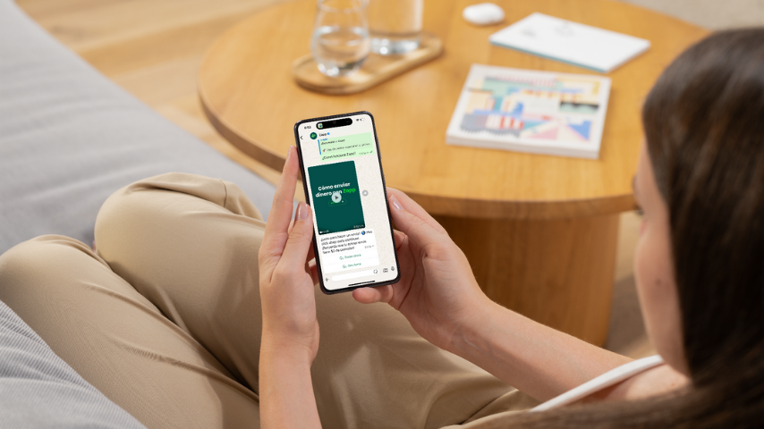 Mujer sentada en un sofá usando su teléfono móvil para ver instrucciones sobre cómo depositar dinero en la cuenta bancaria de otra persona, con una mesa de madera y documentos al fondo.