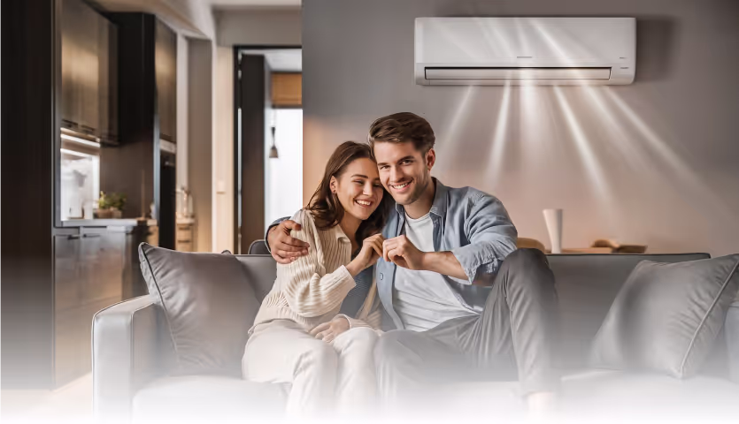 a smiling couple sitting on a couch in a living room, with a wall-mounted air conditioning unit blowing air above them