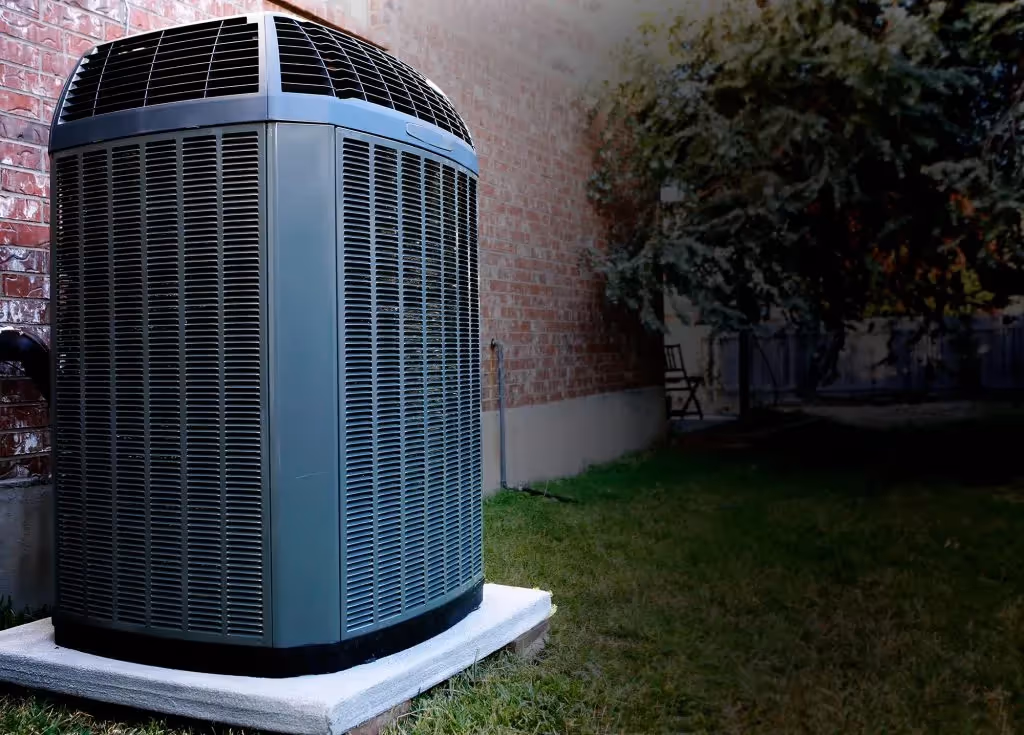 An outdoor air conditioning unit, gray and cylindrical with vertical grates, sits on a concrete slab next to a brick wall. The unit is placed on a green lawn with a blurred background showing a tree and a small chair. The lighting suggests it may be dusk or nighttime.