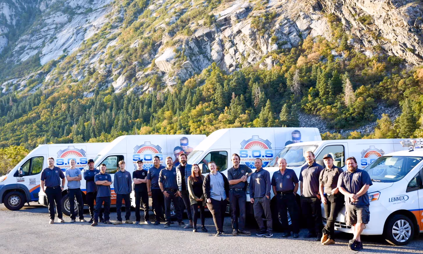  A large group of male and female HVAC technicians, dressed in professional uniforms with company logos, are standing in a long line in front of a fleet of white service vans. The vans are branded with "S.O.S. HEATING & COOLING" and feature superhero-themed graphics. They are positioned outdoors in what appears to be a parking area at the base of a large, rugged mountain covered with sparse trees and vegetation. The group is smiling and posing for the camera under natural light.