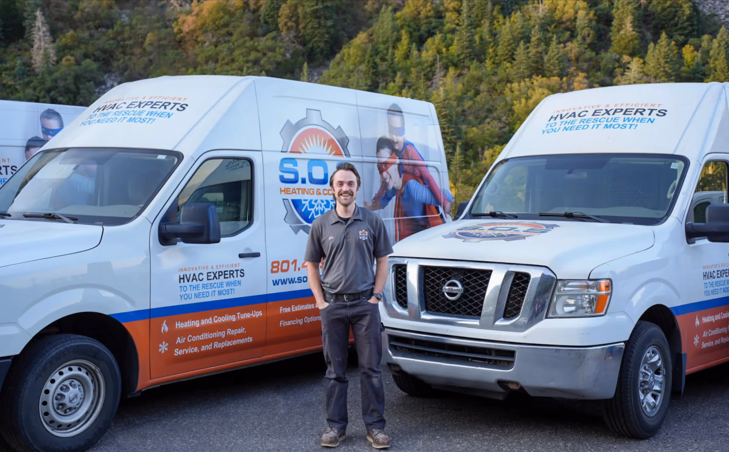 A man wearing a grey polo shirt and jeans stands between two white company vans. The vans are branded with a logo that says "S.O. Heating & Cooling" and "HVAC EXPERTS TO THE RESCUE WHEN YOU NEED IT MOST!". The branding also includes a red and blue graphic of a superhero. The man is smiling at the camera with his hands in his pockets. The background shows trees and a mountain, suggesting an outdoor or rural setting.