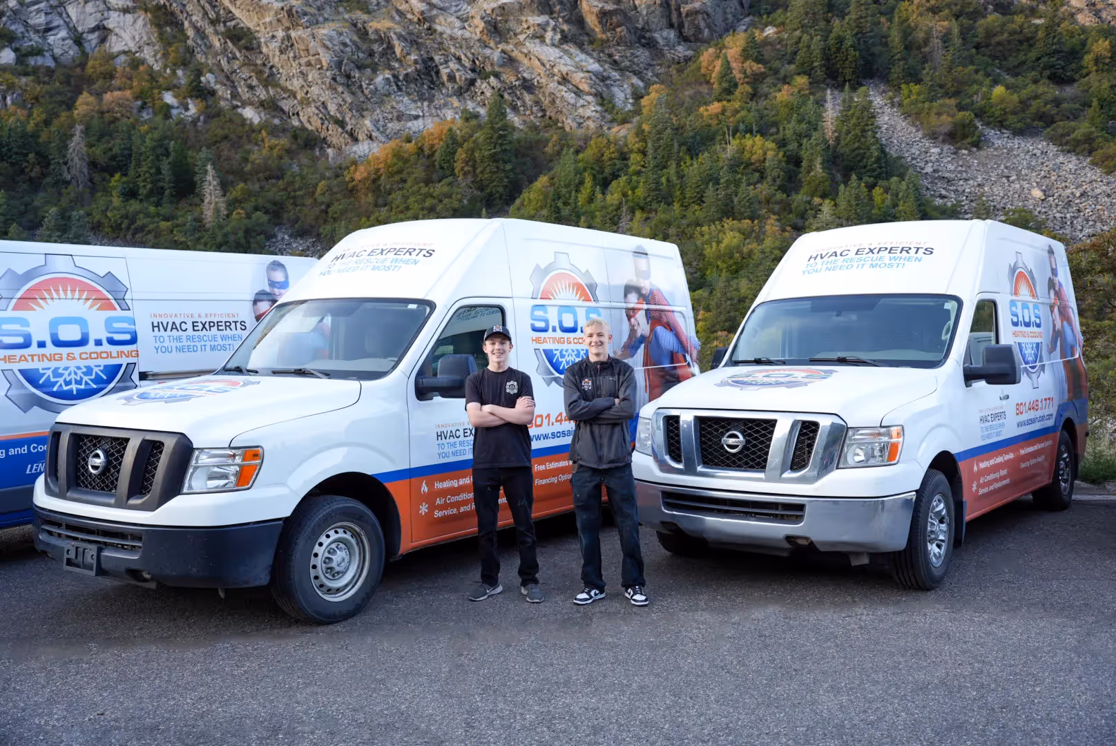  Two male HVAC technicians stand side-by-side, smiling at the camera, in front of two white service vans branded "S.O.S. HEATING & COOLING." The technician on the left is wearing a black t-shirt with a logo and black pants, while the one on the right is wearing a dark zip-up jacket over a shirt and dark pants. Both have their arms crossed. The vans feature superhero-themed graphics and text like "HVAC EXPERTS TO THE RESCUE WHEN YOU NEED IT MOST!" A tree-covered mountain or hillside is visible in the background under a clear sky.