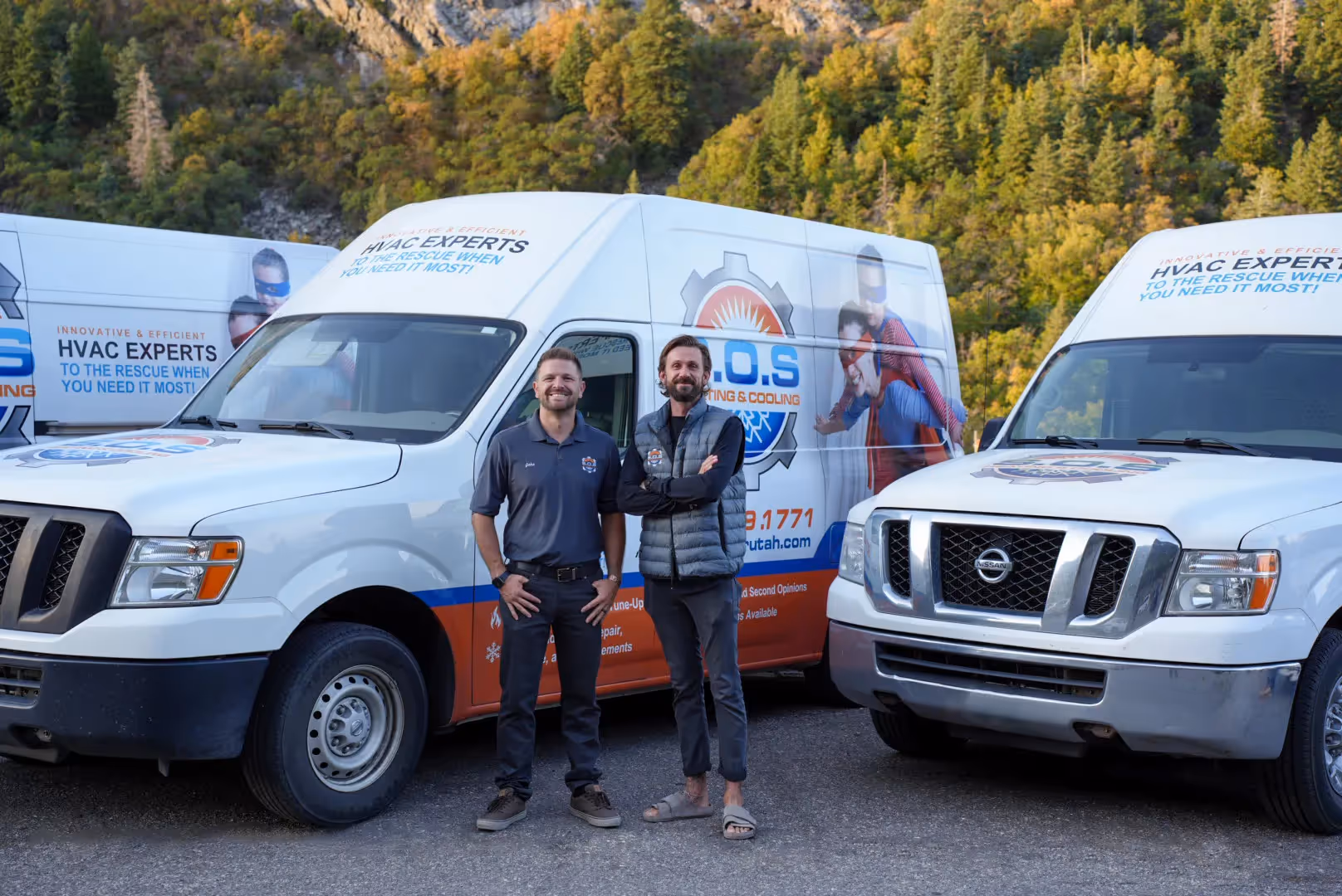  Two male HVAC technicians stand side-by-side, smiling at the camera, in front of two white service vans branded "S.O.S. HEATING & COOLING." The technician on the left wears a dark gray polo shirt with a logo and dark pants. The technician on the right wears a dark long-sleeved shirt, a gray and black puffer vest, and dark pants, with his arms crossed. The vans feature superhero-themed graphics and text like "INNOVATIVE & EFFICIENT HVAC EXPERTS TO THE RESCUE WHEN YOU NEED IT MOST!" A tree-covered mountain or hillside is visible in the background under a clear sky.