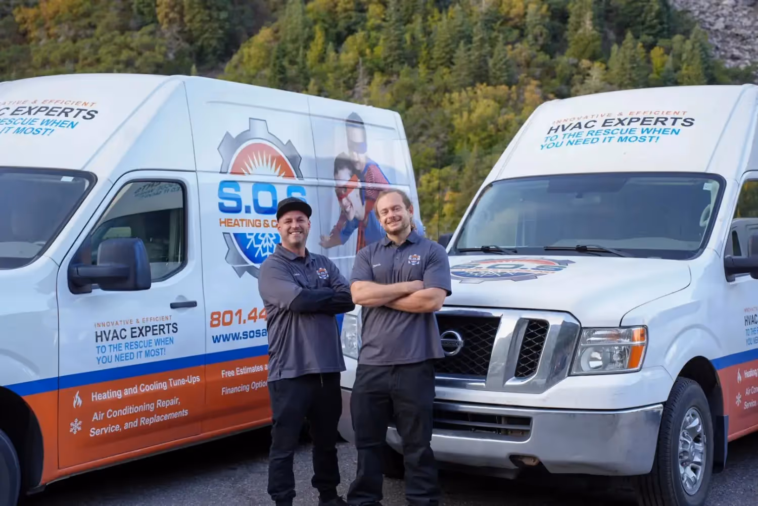  Two male HVAC technicians stand side-by-side with their arms crossed, smiling at the camera, in front of two white service vans branded "S.O.S. HEATING & COOLING." The technician on the left is wearing a dark gray polo shirt, a baseball cap, and dark pants. The technician on the right, with longer hair and a beard, is also wearing a dark gray polo shirt and dark pants. The vans feature superhero-themed graphics and text like "INNOVATIVE & EFFICIENT HVAC EXPERTS TO THE RESCUE WHEN YOU NEED IT MOST!" A tree-covered mountain or hillside is visible in the background under a clear sky.