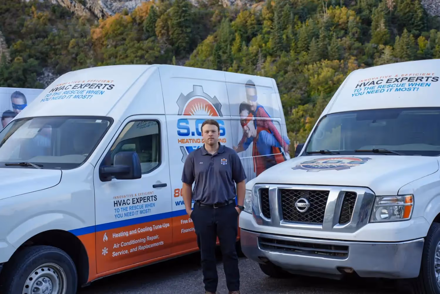 A male HVAC technician, smiling and with his arms crossed, stands in front of two white service vans. He is wearing a gray polo shirt with a logo, dark pants, and black athletic shoes. The vans are branded with "S.O.S. HEATING & COOLING" and feature superhero-themed graphics and text like "INNOVATIVE & EFFICIENT HVAC EXPERTS TO THE RESCUE WHEN YOU NEED IT MOST!" A tree-covered mountain or hillside is visible in the background under a clear sky.