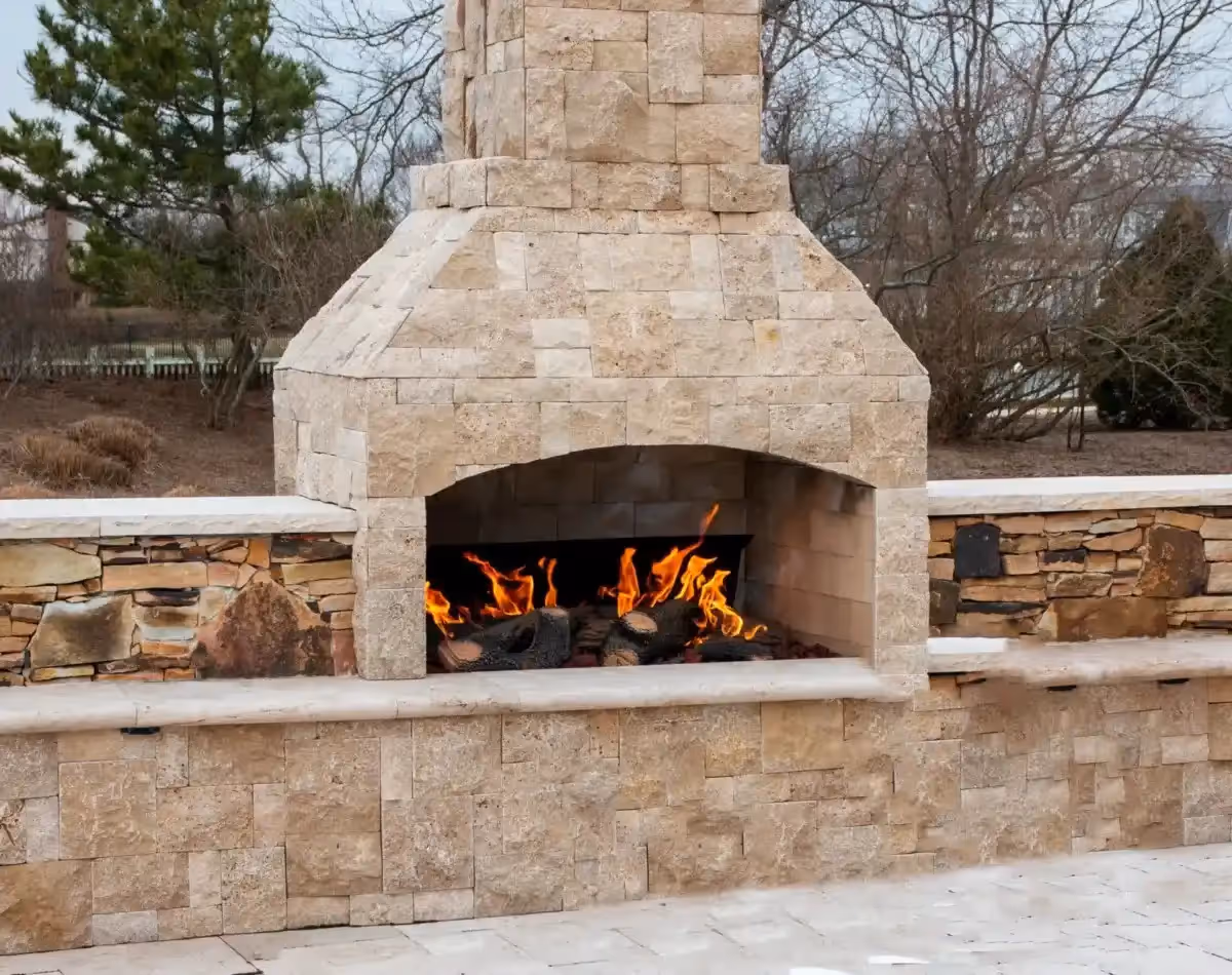 An outdoor fireplace constructed from light-colored, possibly travertine, stone blocks, with a fire burning inside its arched opening. The fireplace features a tiered chimney and is built into a low wall of the same stone. In front of the fireplace, there's a paved patio area. The background shows bare trees and some greenery under an overcast sky.