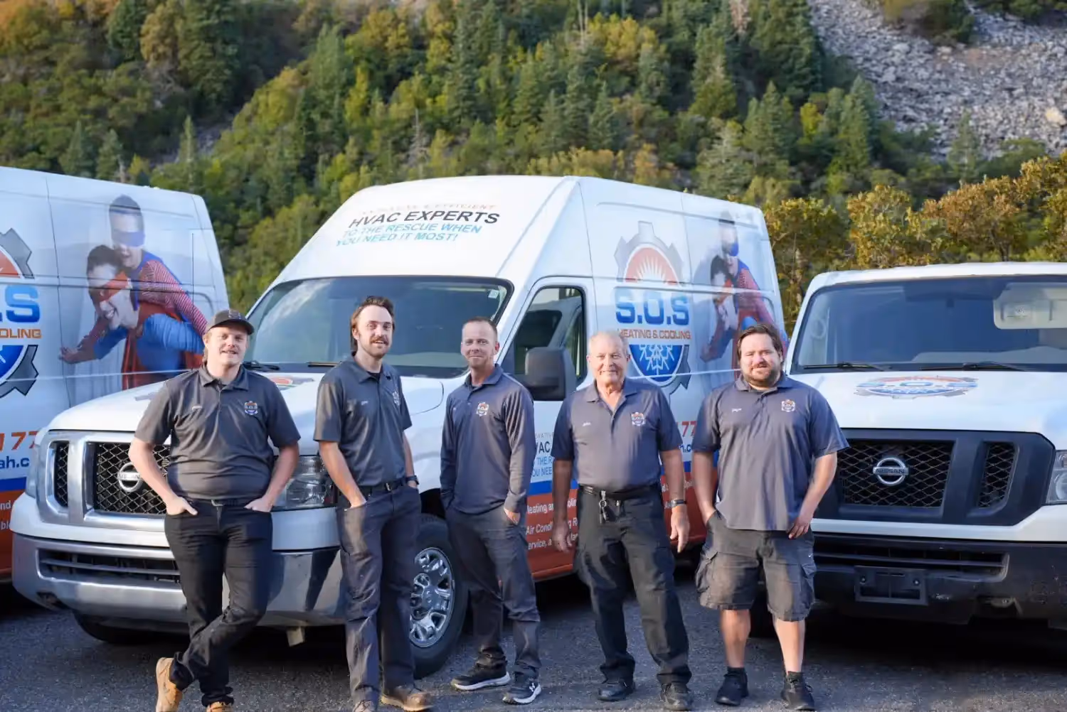 A group of five male HVAC service technicians, dressed in dark gray shirts with logos and various pants (jeans, shorts, cargo pants), are standing outside in front of three white service vans. The vans are branded with "S.O.S. HEATING & COOLING" and feature graphics of superheroes. A tree-covered mountain or hillside forms the backdrop under a clear sky. The technicians are smiling and posing for the camera.