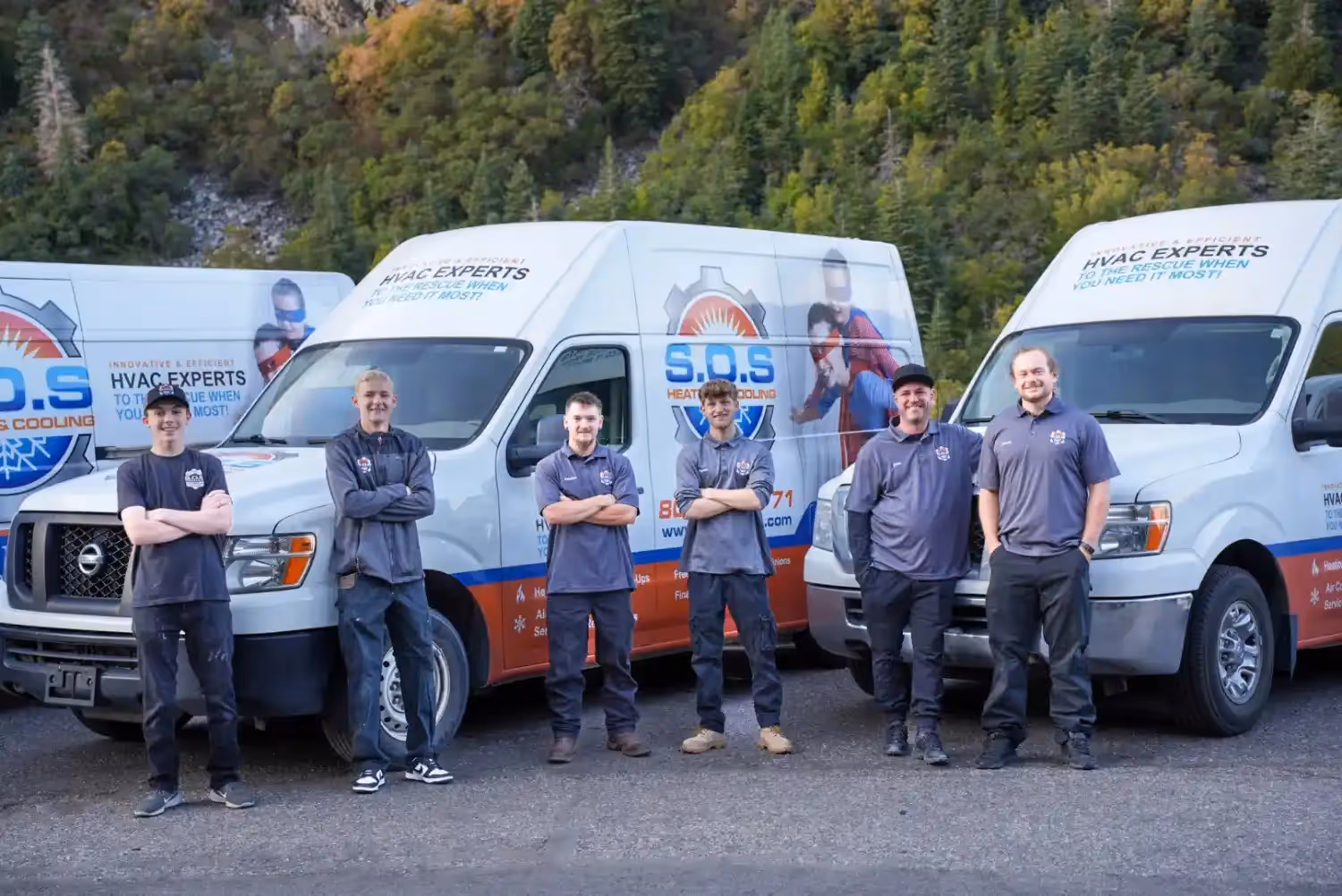 A group of six male HVAC technicians in various grey and blue work uniforms stand smiling in front of three white service vans with "S.O.S. Heating & Cooling" branding, with mountains and trees in the background.