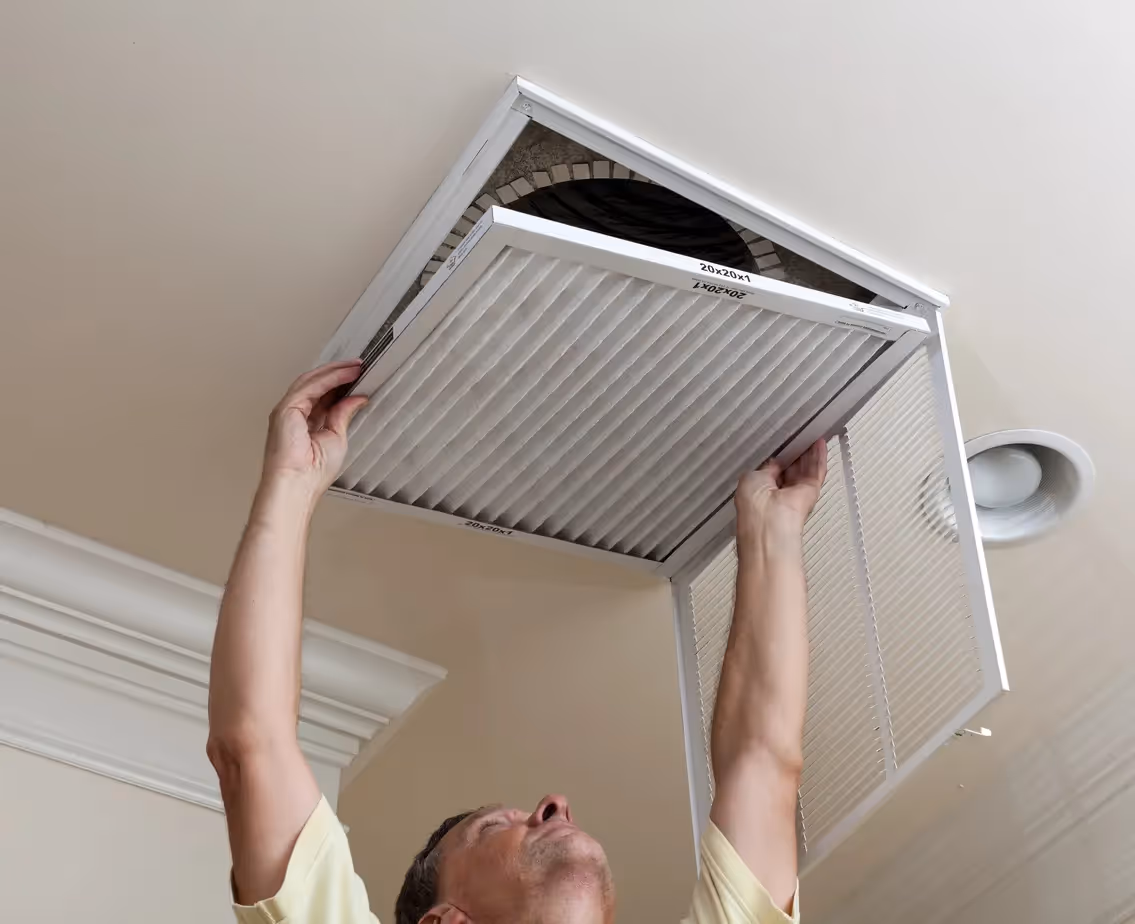  A person, viewed from a low angle, is reaching up to remove a rectangular, pleated white air filter from a ceiling vent. Their hands are holding the edges of the filter and the vent cover. The ceiling is light-colored, and a circular light fixture is visible to the right of the vent. The inside of the vent shows some ductwork or a fan.