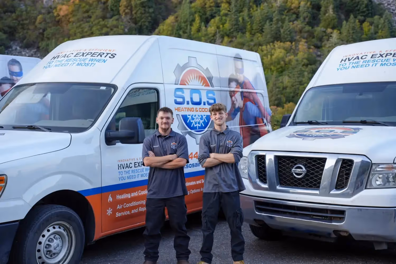 Two smiling male technicians stand in front of three white HVAC service vans, with mountains and trees in the background.