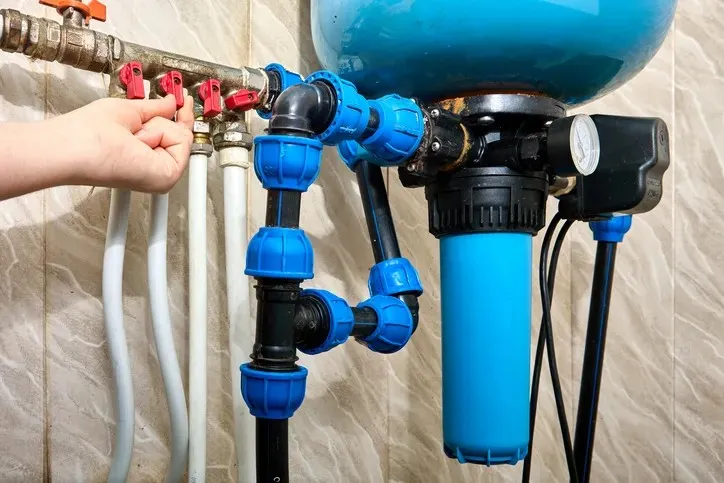 A hand turning red valves on a complex blue and copper pipe system with a large blue water filter