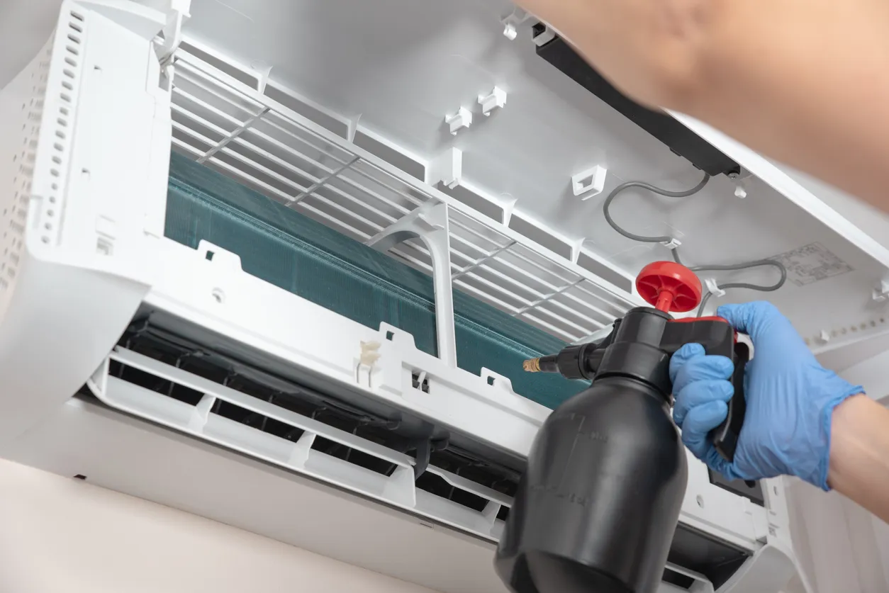 A person wearing blue gloves sprays a cleaning solution into the open internal components of a white wall-mounted air conditioning unit.